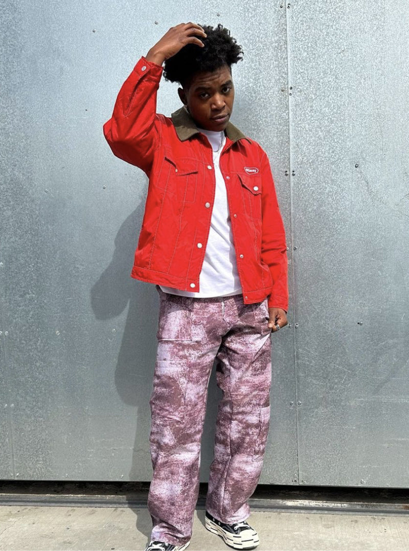 Masc person posing and wearing bright red  jean jacket with a white undershirt and purple patterned cargo pants