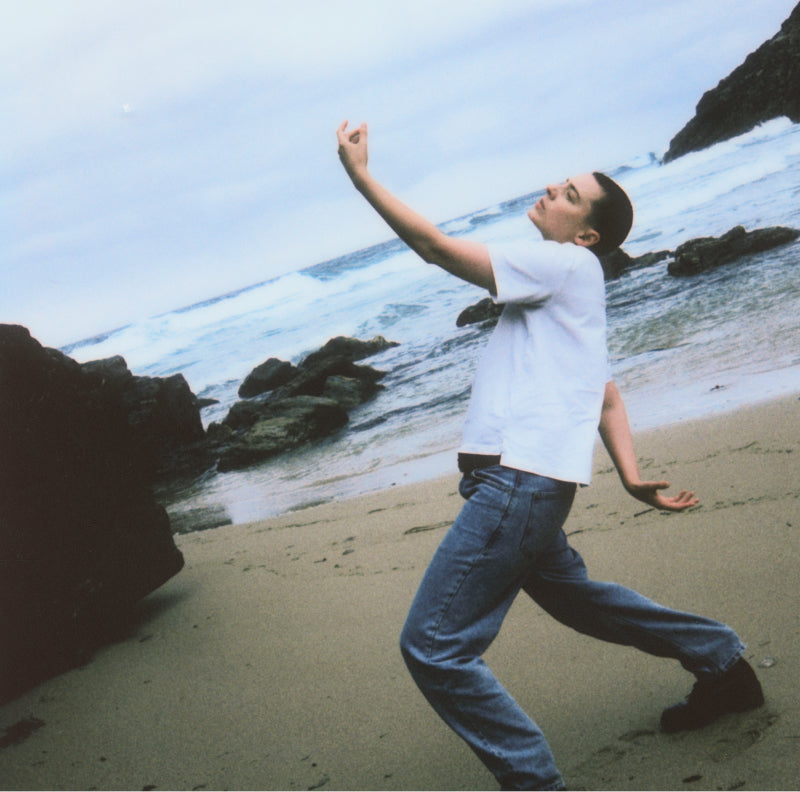 Orion Sacs, figure in white tee and jeans dancing on a beach