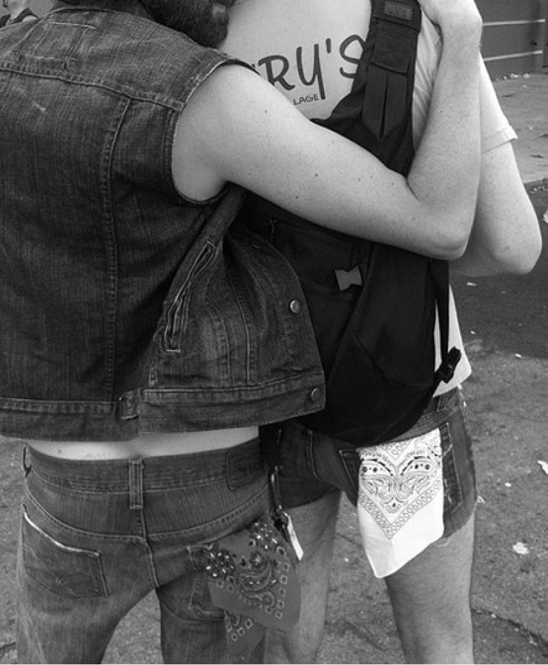 Black and White photo of two men who are standing leaning on each other with their backs to the camera to show handkerchiefs in their back pockets as a symbol of their queerness.