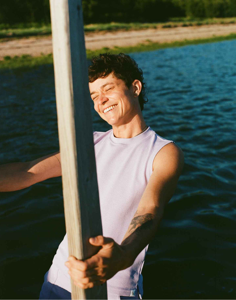 Transmasc person smiling and wearing lilac romeo swim tank, outside by a lake