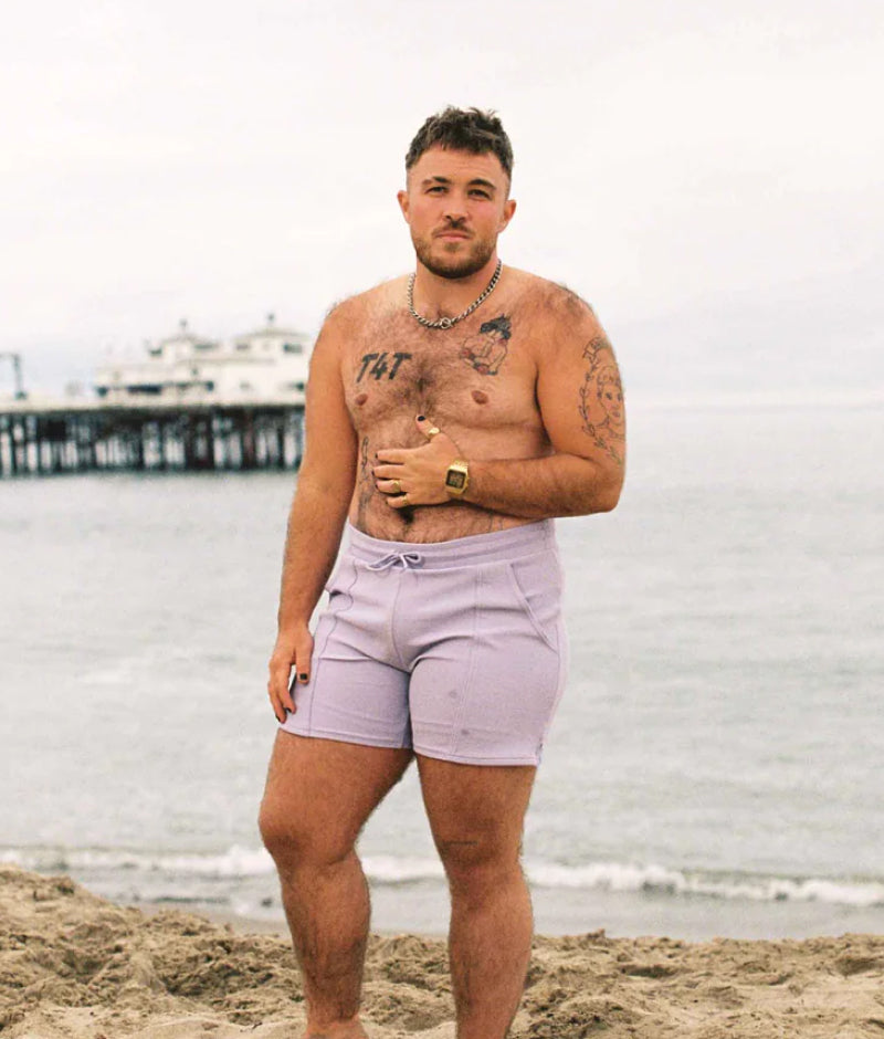 Trans man shirtless in lilac Elliot swimtrunks, standing at a beach