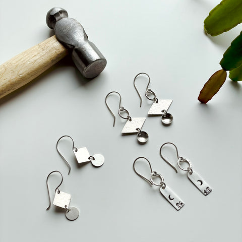 Three pairs of silver drop earrings made from geometric shapes with hammered textures and stamped symbols with a hammer in the background