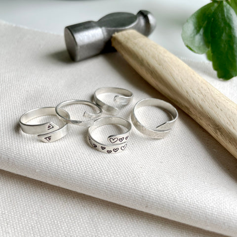 A collection of silver wrap rings decorated with hammered textures and stamped symbols and hearts with a jewellers hammer and plant leaves in the background