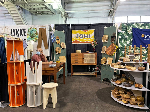 Face on perspective of JOHI's West Coast Craft booth in San Francisco in September 2021. A quilted yellow/rust/white sign with gray letters reading "JOHI" hangs at the upper center on a black curtain backdrop. A wood & woven danish cord console sits in the middle back, flanked by two green painted peg boards on either side. Product is displayed on the peg boards, like bodysurf handplanes and kitchen trivets. Founder Raja sits on the left at a hardwood desk. In the front left of the photo are a display of stools. At the front right of the photo is a display of small geo block planters and geometric lamps.