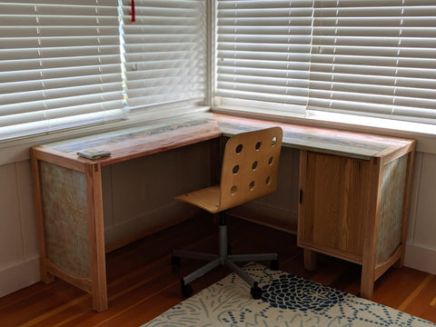 Looking into the corner of a room, the blinds on adjacent windows are pulled down to shade the interior. The new Kalakoa Desk is paired with a bent plywood office chair on wheels. A floral blue and white rug extends from the bottom right of the photo.