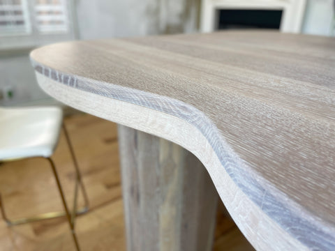Photo focuses on the curving edges of a custom designed white oak table by JOHI. The white oak is stained white, with wood grain showing through.