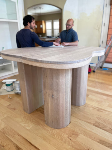 Two men stand behind the curving, custom designed white oak table. Its pillar legs rise tall, so the table is at a standing, bar height. The men are contemplating ideas over a sketchbook.