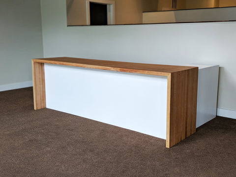 The entry desk at this commercial office is made of red oak with a waterfall style. A white panel underneath the table hides activity and clutter behind the desk, and a white cabinet is inset in a perpendicular orientation to the desk. The office has a brown carpet and creamy white walls.