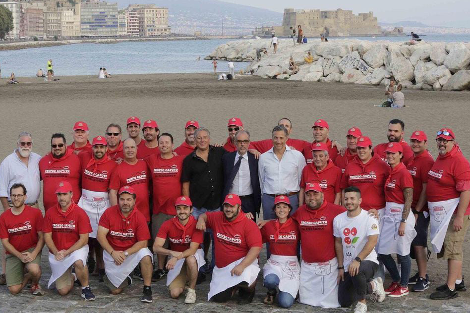 A group photo of 2 dozen pizza makers posing with executives of Caputo and Orlando Food Sales on a beach in Italy