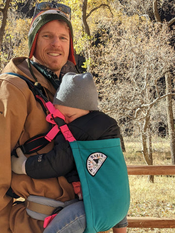 A man carrying a child on his front with a Trail Magik carrier attached to his hiking pack while hiking
