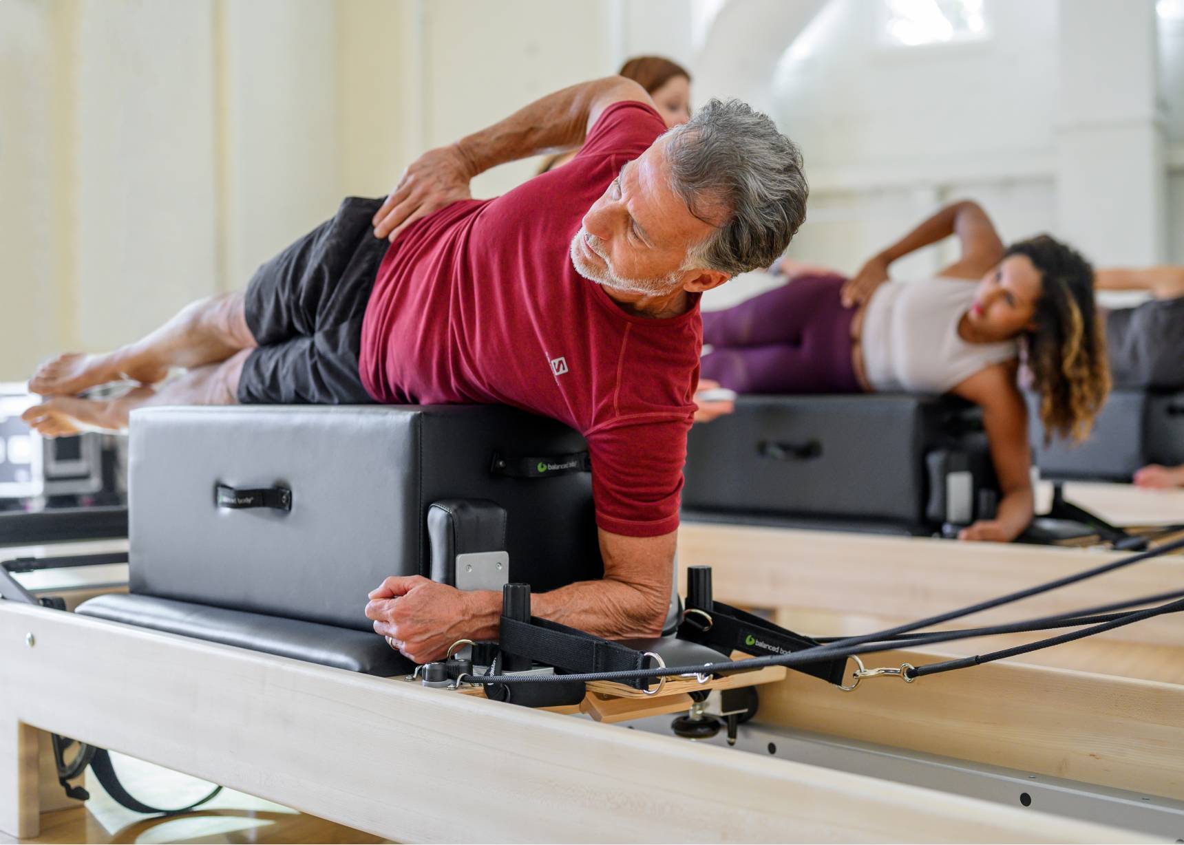 Man laying across a sitting box that is on top of a Pilates Reformer with other students in the background