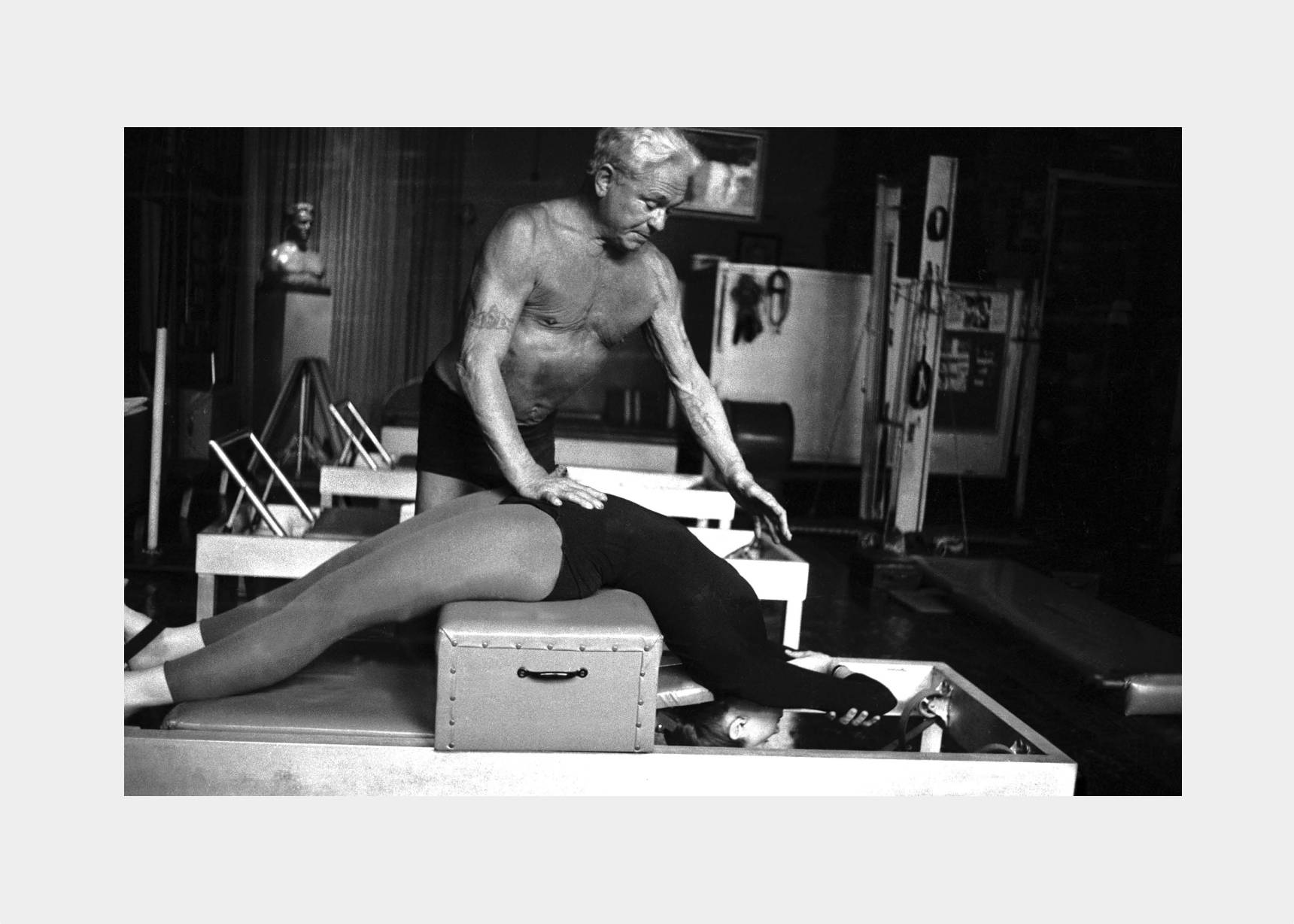 Vintage photograph of a student guided by Joseph Pilates on a sitting box and reformer. 