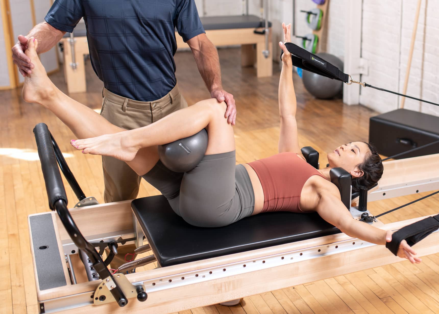 A physical therapist reaches down to adjust the knee position of a client laying on a pilates reformer