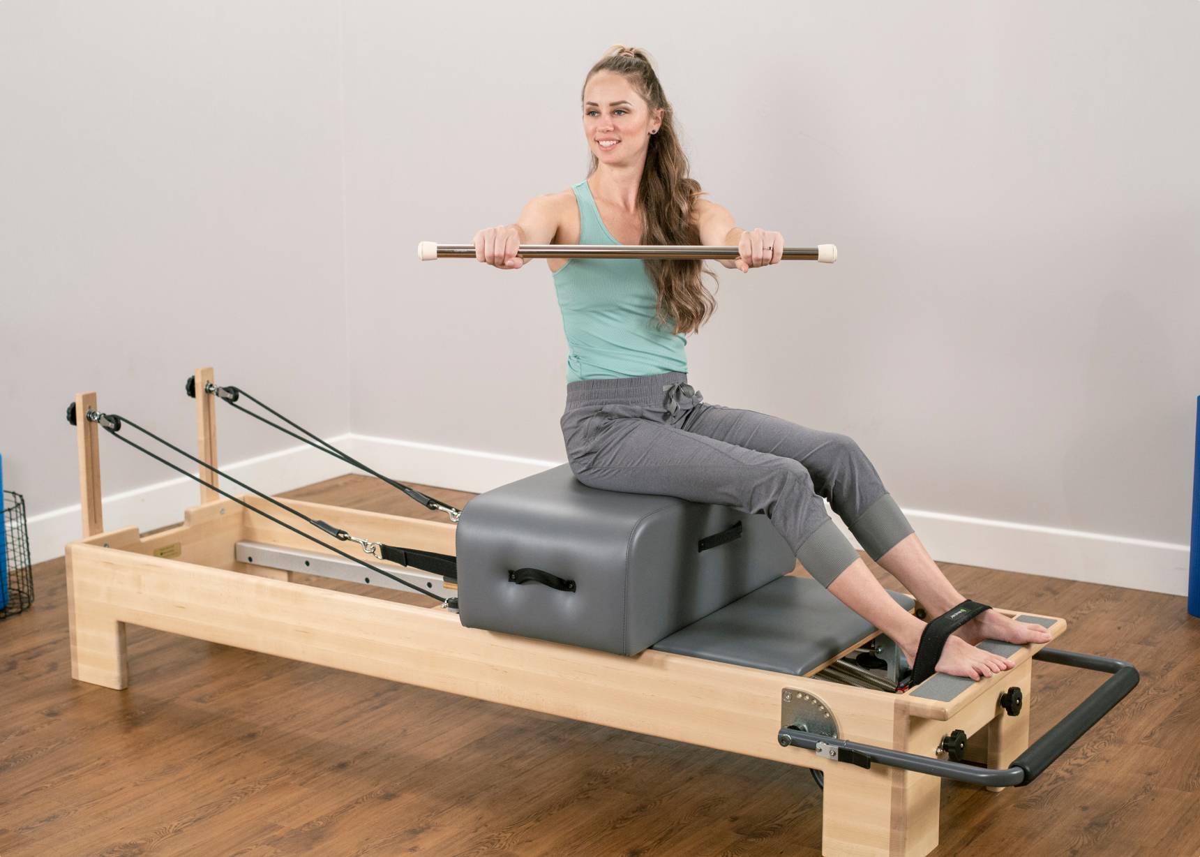 A woman exercising on a contour sitting box, using a maple dowel and attached Pilates foot straps.