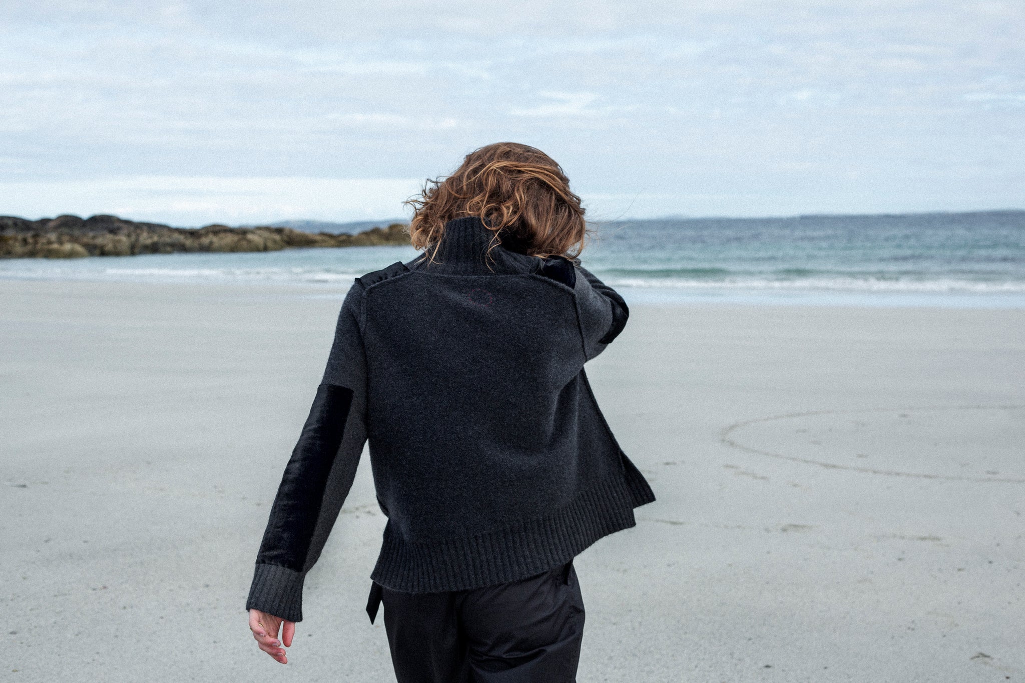 White woman with wavy brown hair walks away from camera on a fine sand beach in Connemara, West of Ireland. She is wearing Irish designed luxury cashmere sweater in a Charcoal mélange colour from Loro Piana. It has shoulder epaulettes and long patches on the lower sleeve made from Black velvet.