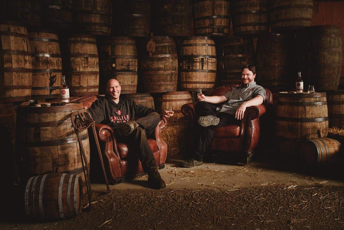 Owners of Shelter Point Distillery sitting on arm chairs in their distillery surrounded by whisky barrels and whisky glasses in hand