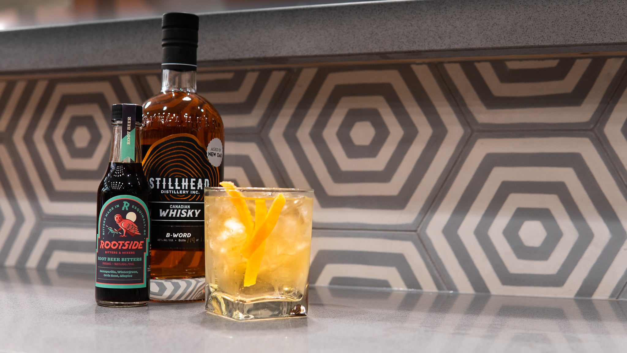 Stillhead's BWord whisky and Rootside's root beer bitter bottles sit behind an old fashioned cocktail with orange peels inside