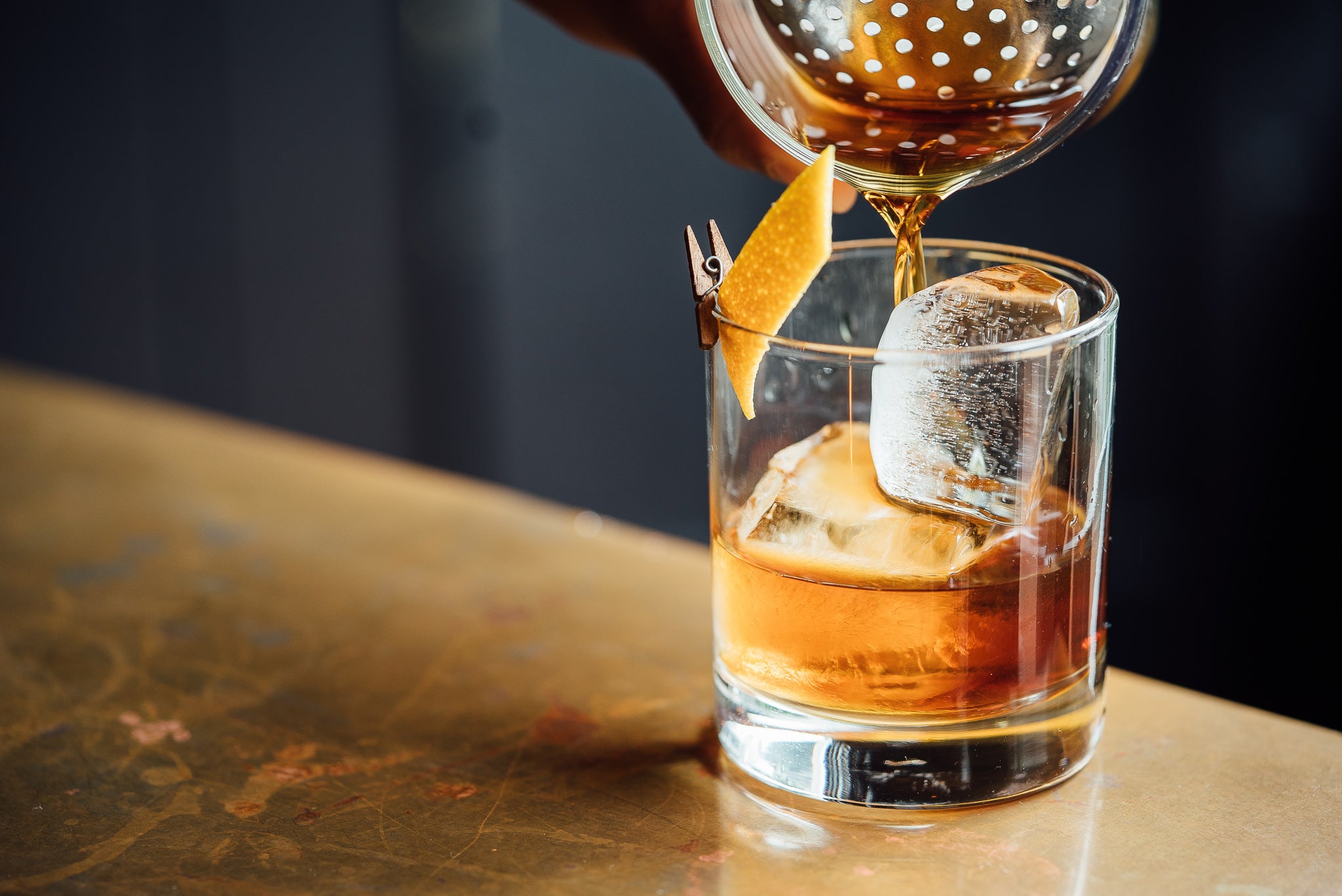 An Old Fashioned being poured into a drinking glass from a shaker with an orange peel garnish