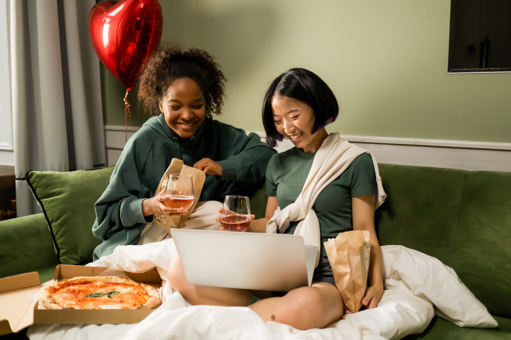 Couple, two women, enjoy a glass of rose together on a couch with a pizza and laptop. A red heart balloon behind them.