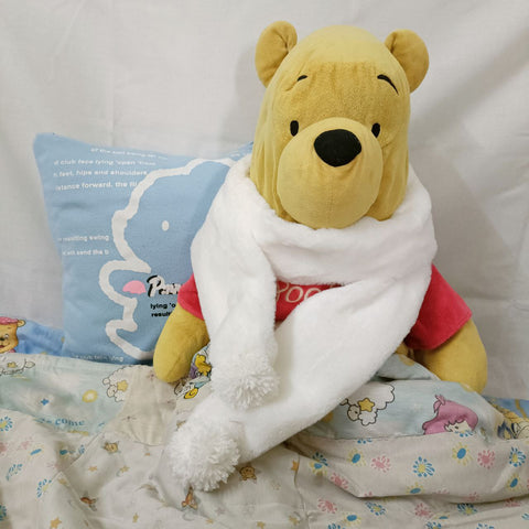 Christmas scarf wrapped around a Pooh bear stuffed toy, cuddled up in a blanket and sitting up against a pillow.