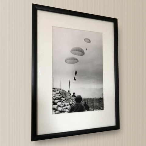 A photo representing a black and white framed photo. It shows paratroopers falling from the sky on the beach while a man, with his back turned to the photographer, watches.
