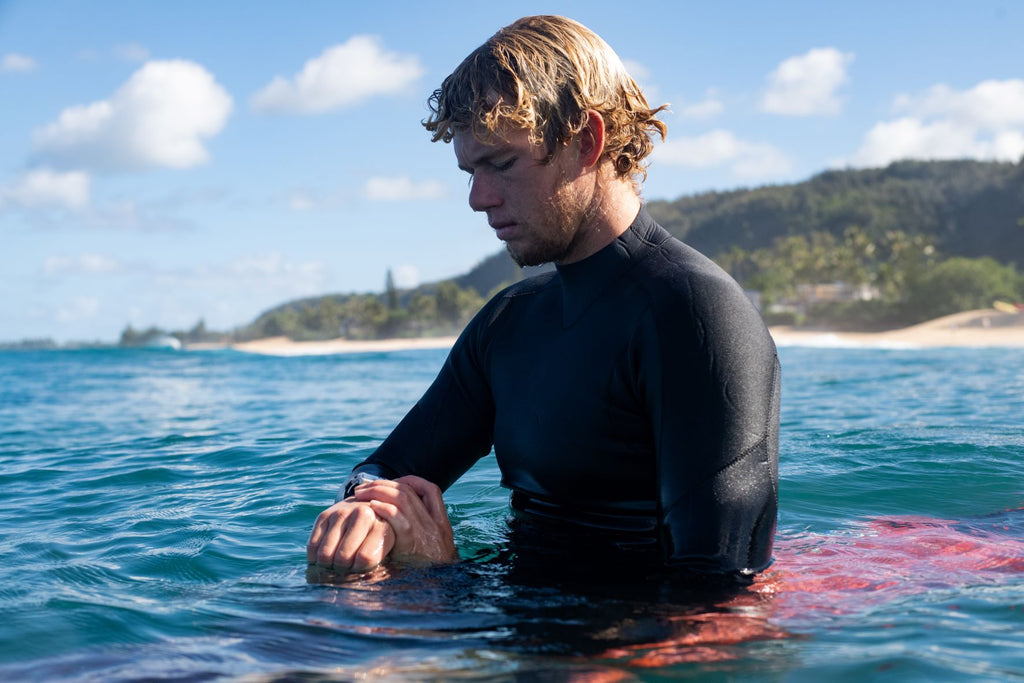 Le surfeur John John Florence est assis dans l'eau et utilise sa montre de surf Nixon