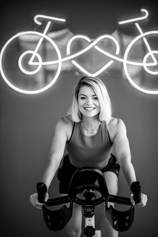 Author of the blog post smiling while sitting on a cycling bike in front of a neon sign with the Cycle of Heart logo