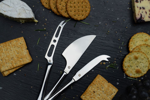 three stainless steel cheese knives on a black slate background with various cheese