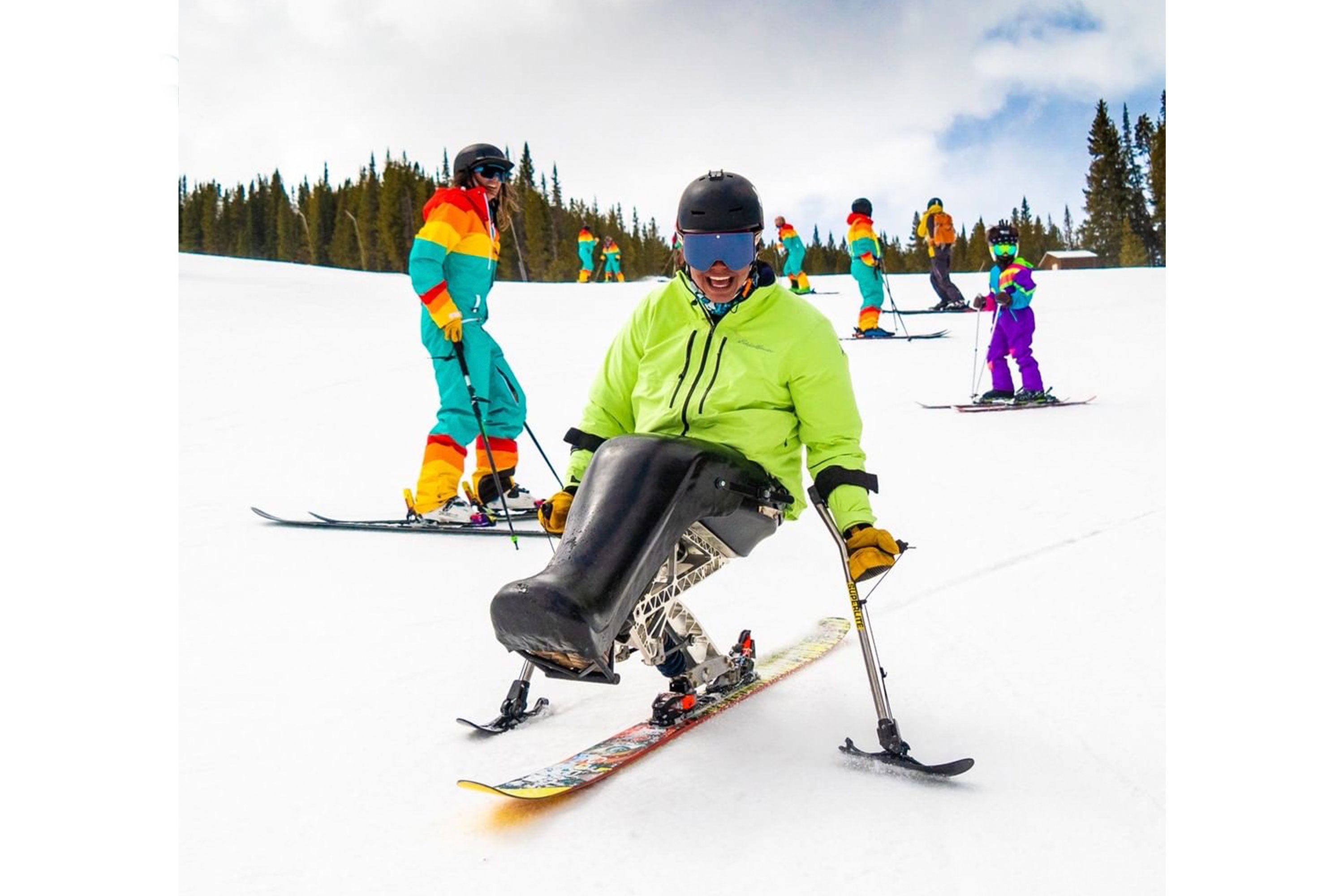 Trevor Kennison sat on his ski set with other skiers behind him on a snowy slope with pine trees in the background