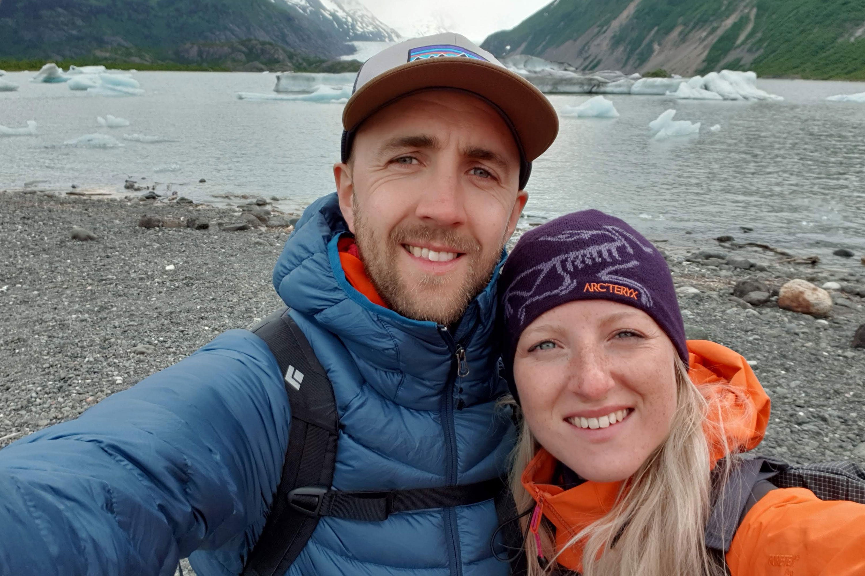 Jade and Scott dressed in cold weather gear, stood in front of Kachemak Bay with blocks of ice and mountains behind