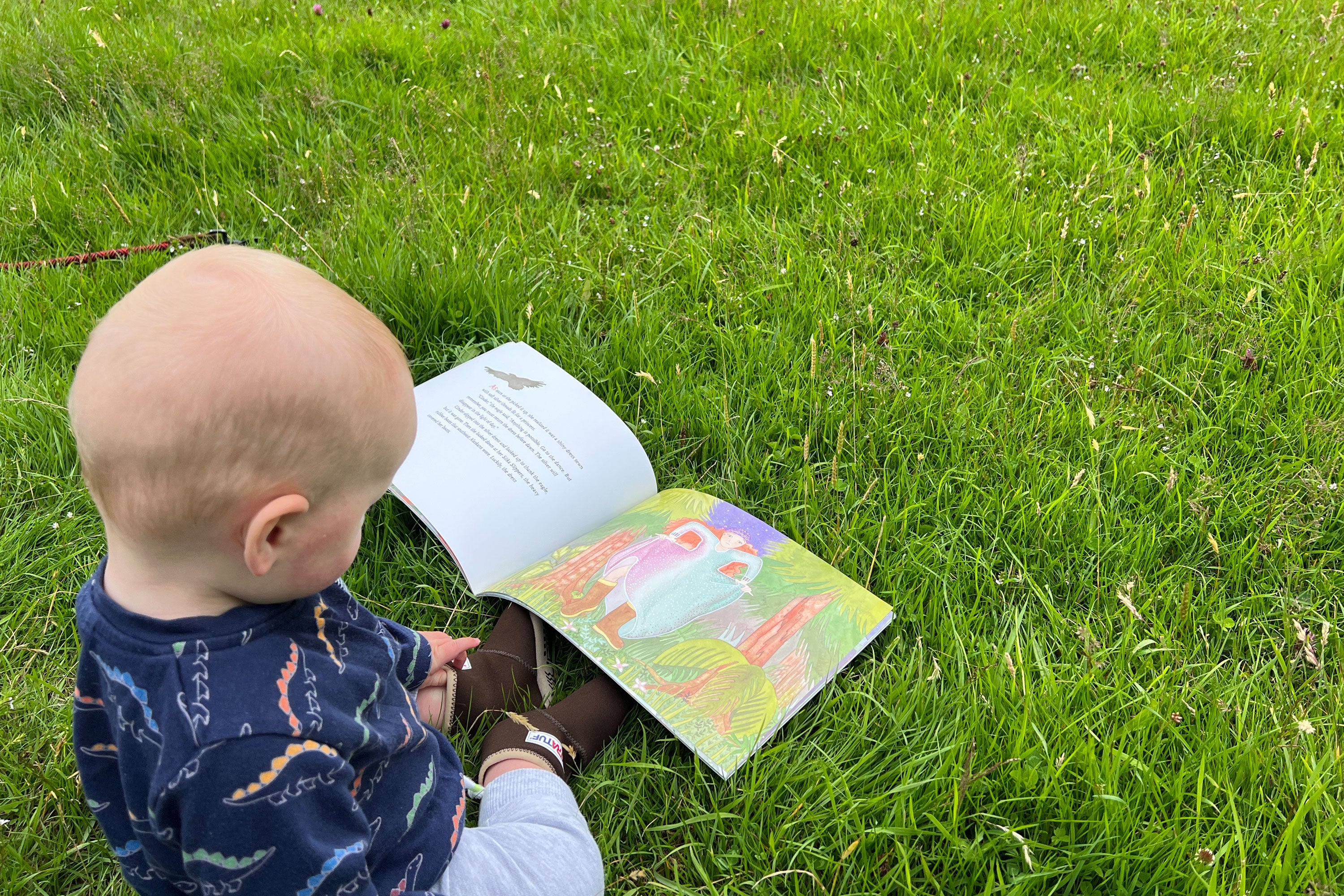 A baby sat on grass wearing a pair of Xtratuf boots, reading a picture book