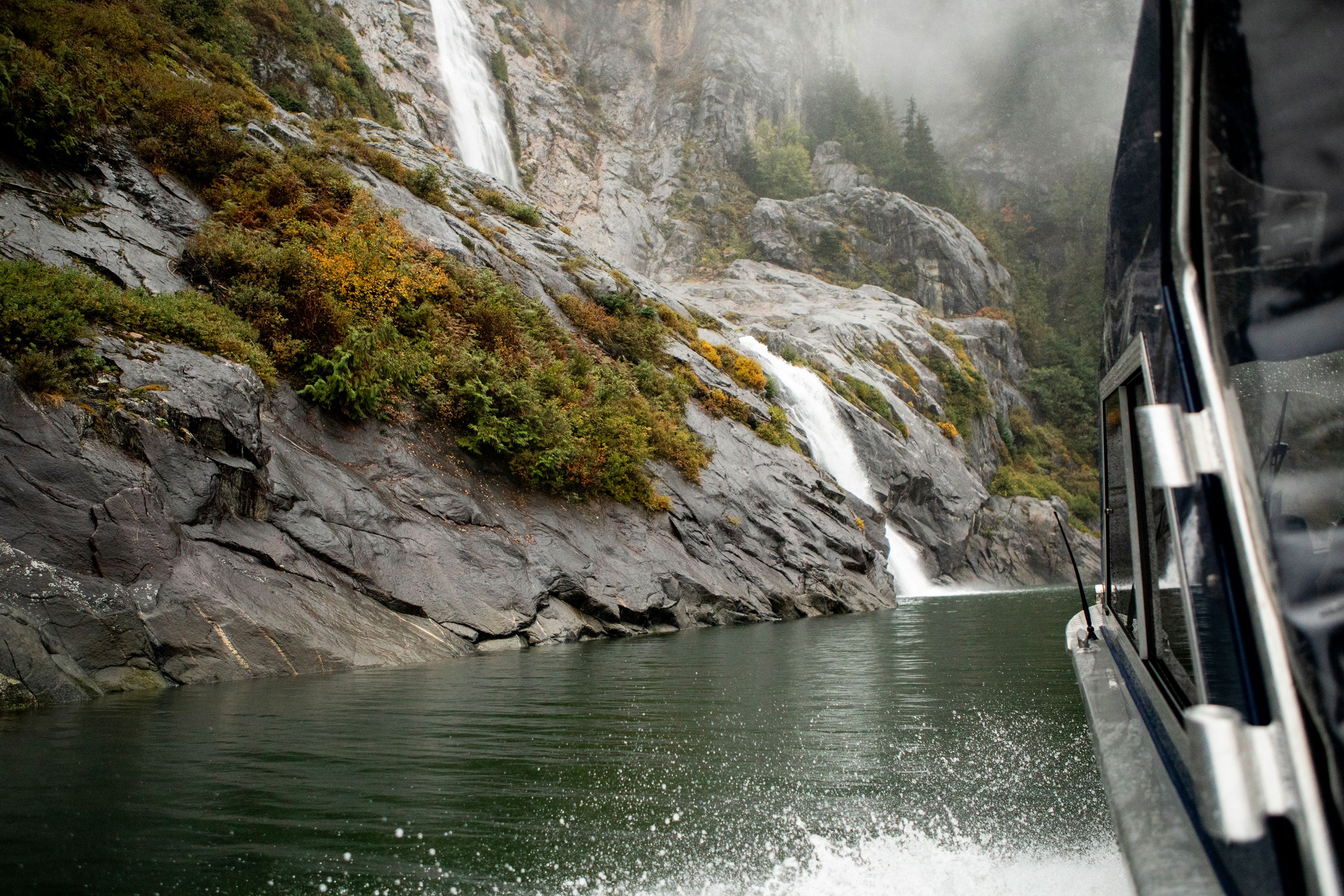 A narrow side view of a boat on water on the right with a mountain face covered in shrubs and pine trees with a waterfall flowing into the sea