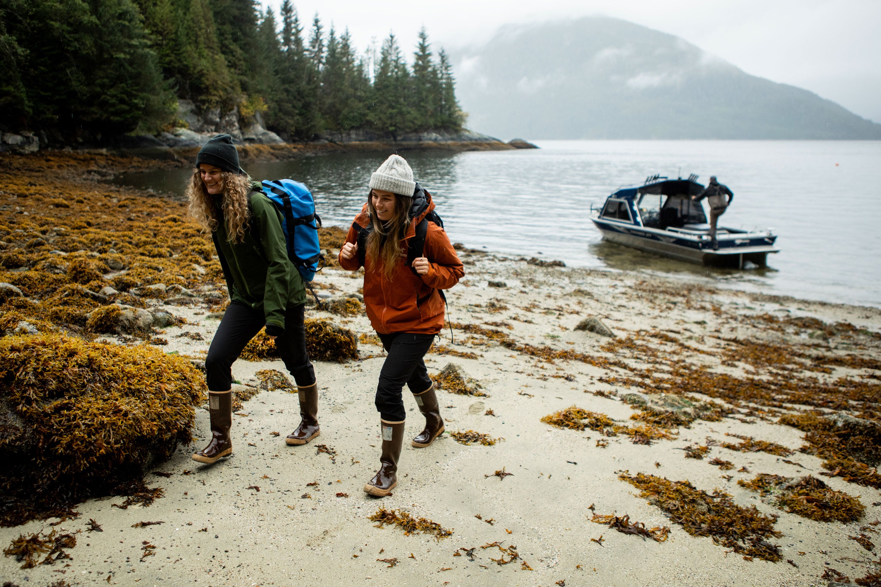 Two women wearing warm & waterproof clothing and Xtratuf Legacy Boots walking along a seaweed covered shore, away from a boat with a man stood on the stern