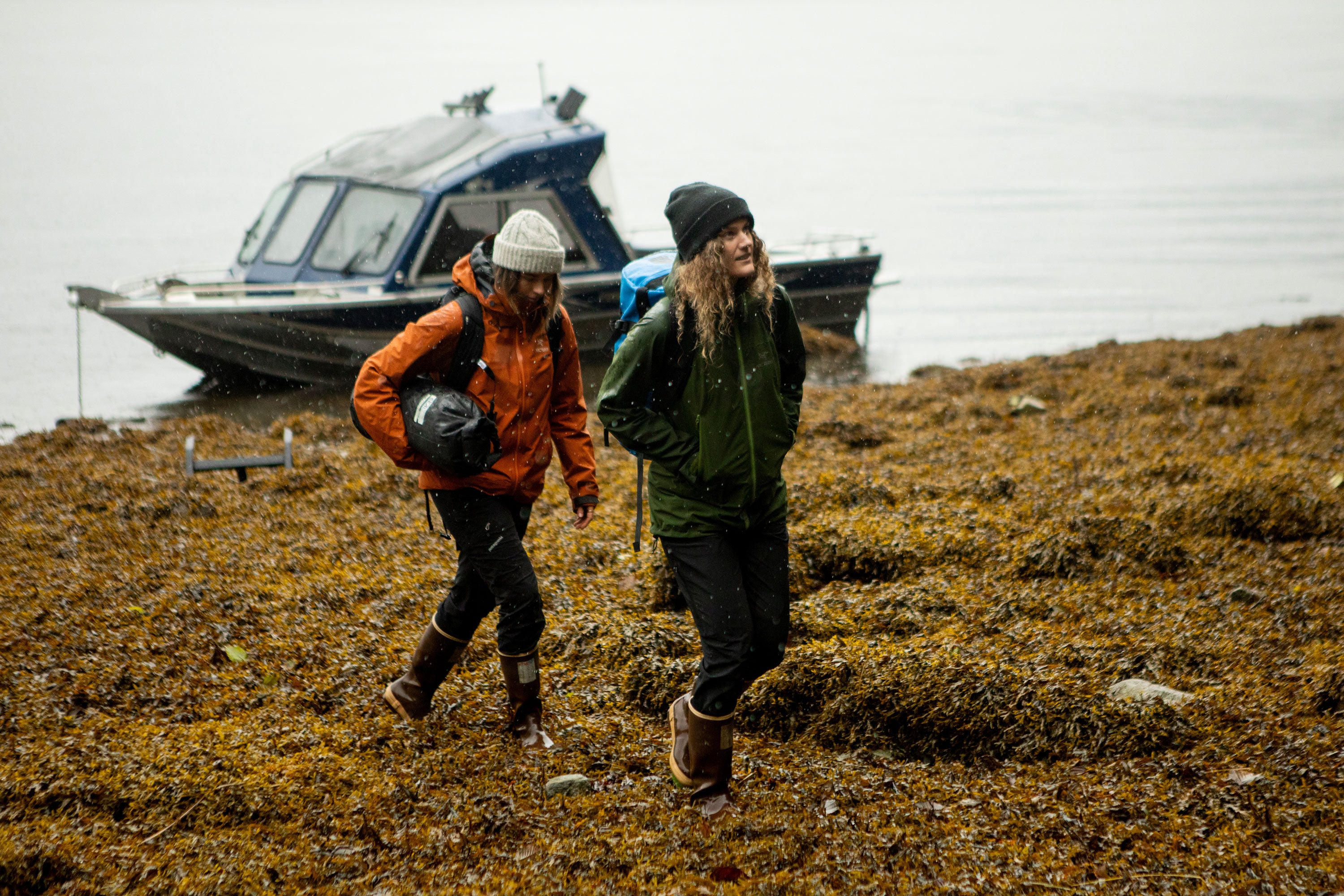 Two women walking along a seaweed covered shore, away from a shored boat. They are both wearing warm, waterproof clothing and Xtratuf Legacy Boots