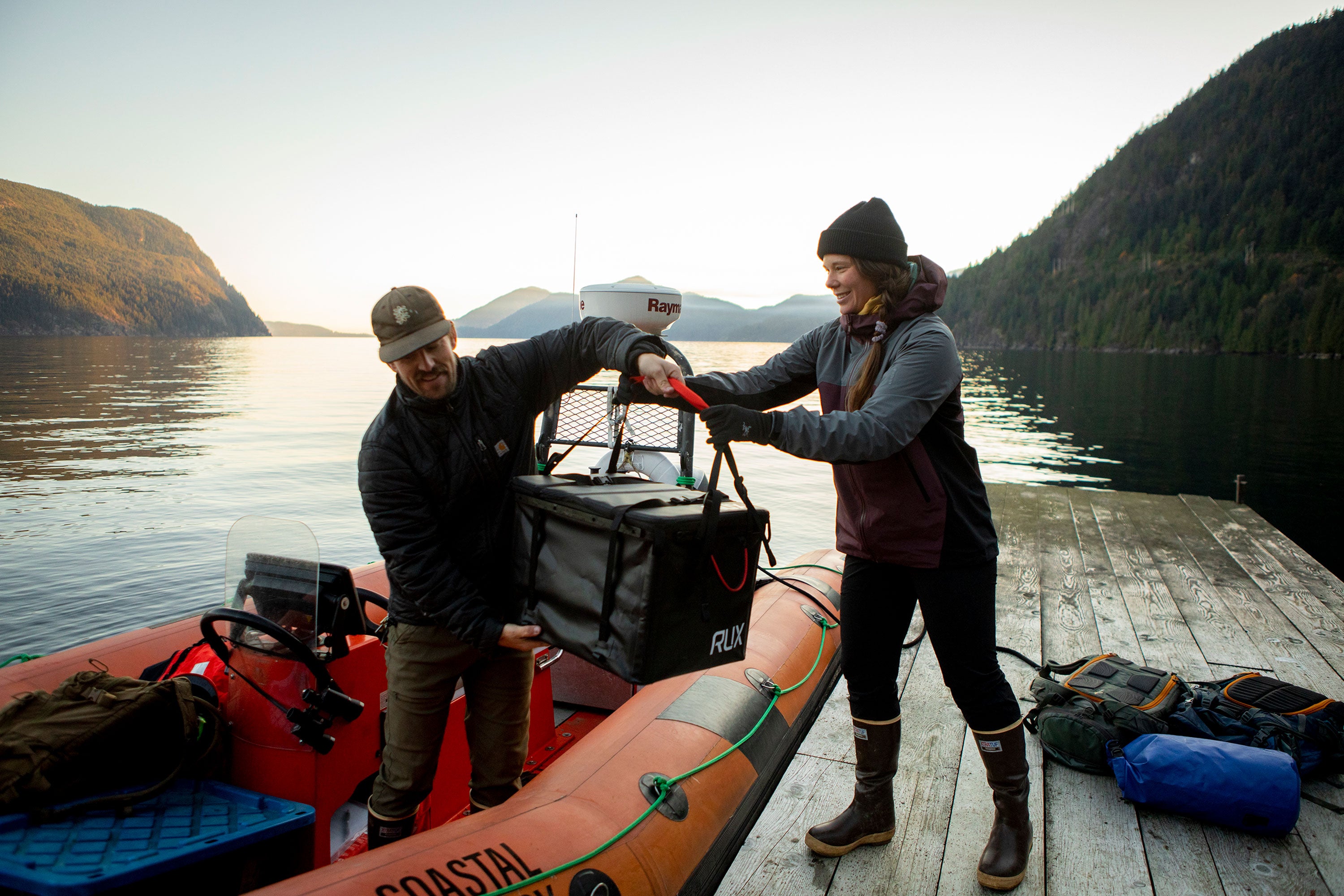 Addison Farr stood on an orange zodiac boat taking a small cargo box from a female colleague stood on a jetty, wearing a pair of brown Xtratuf Legacy boots