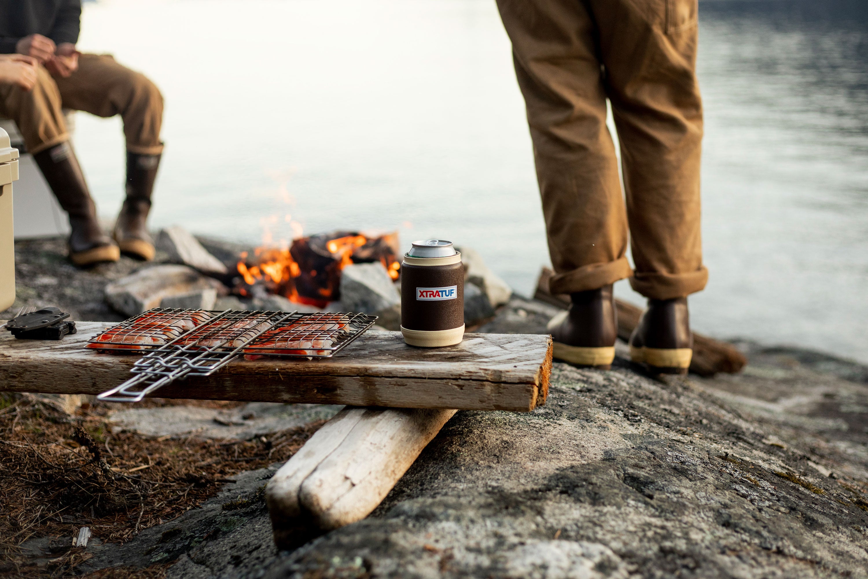 Two people in the background wearing duck brown trousers and Xtratuf Legacy Boots with a bbq fire burning over rocks. In the foreground is a slab of wood with an Xtratuf Can Coozie and fish prepared in a wire roaster