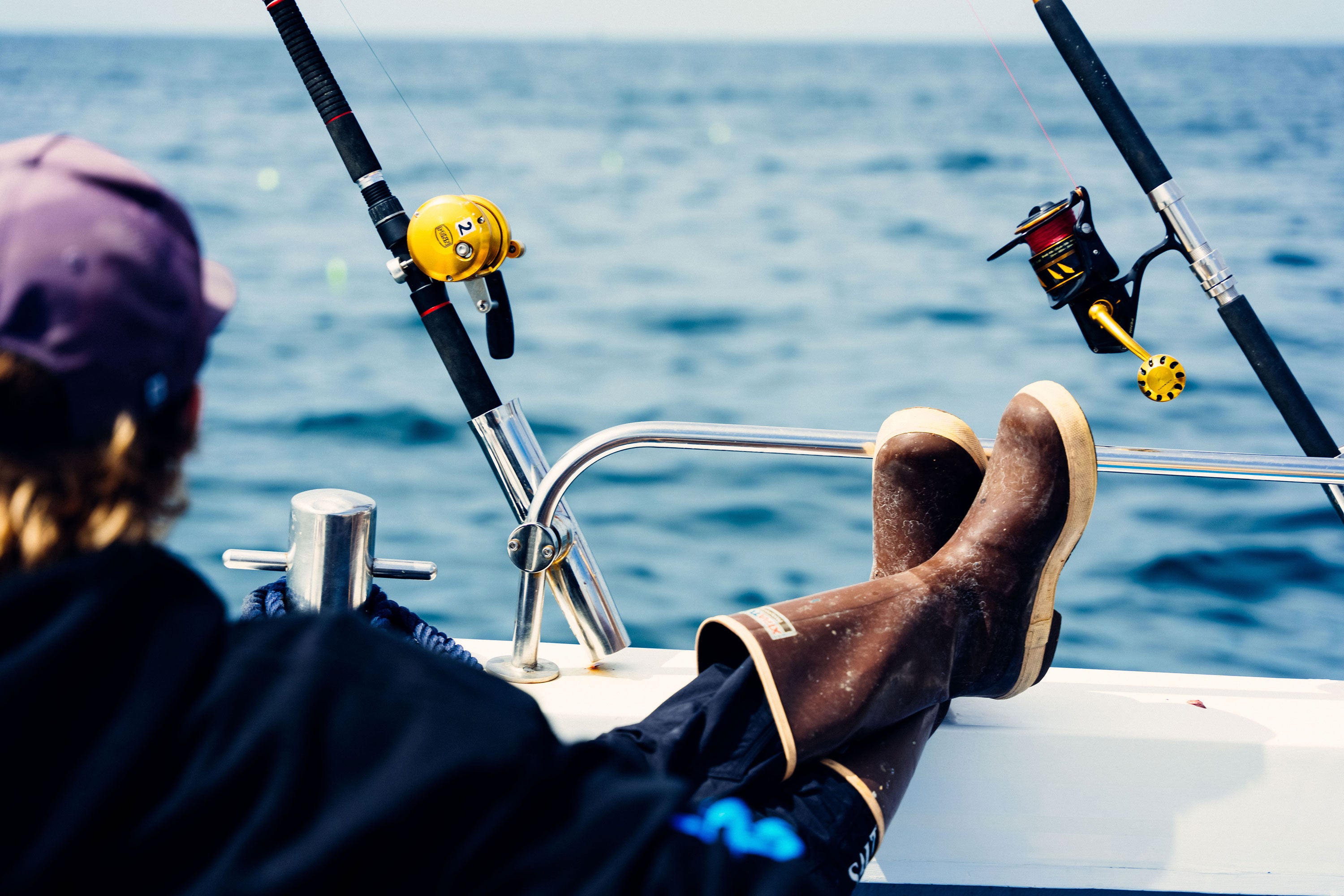 Man sat on a boat with feet up on deck with fishing rods in front. Wearing a pair of Xtratuf Legacy Fishing Boots