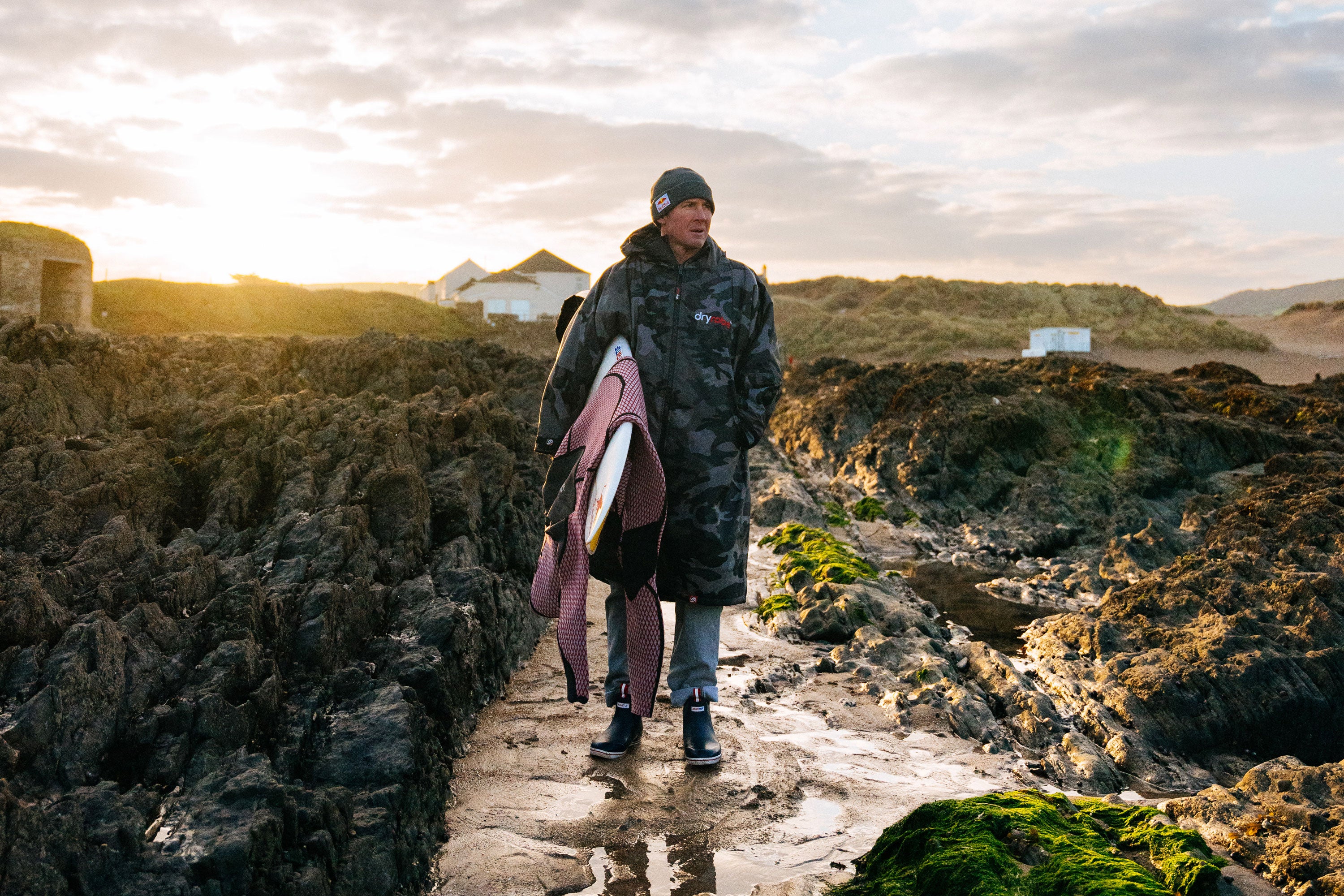 Andrew 'Cotty' Cotton holding a surfboard and wearing a beanie hat, long coat and pair of Xtratuf ankle deck boots whilst stood on wet rocks with the sun rising behind
