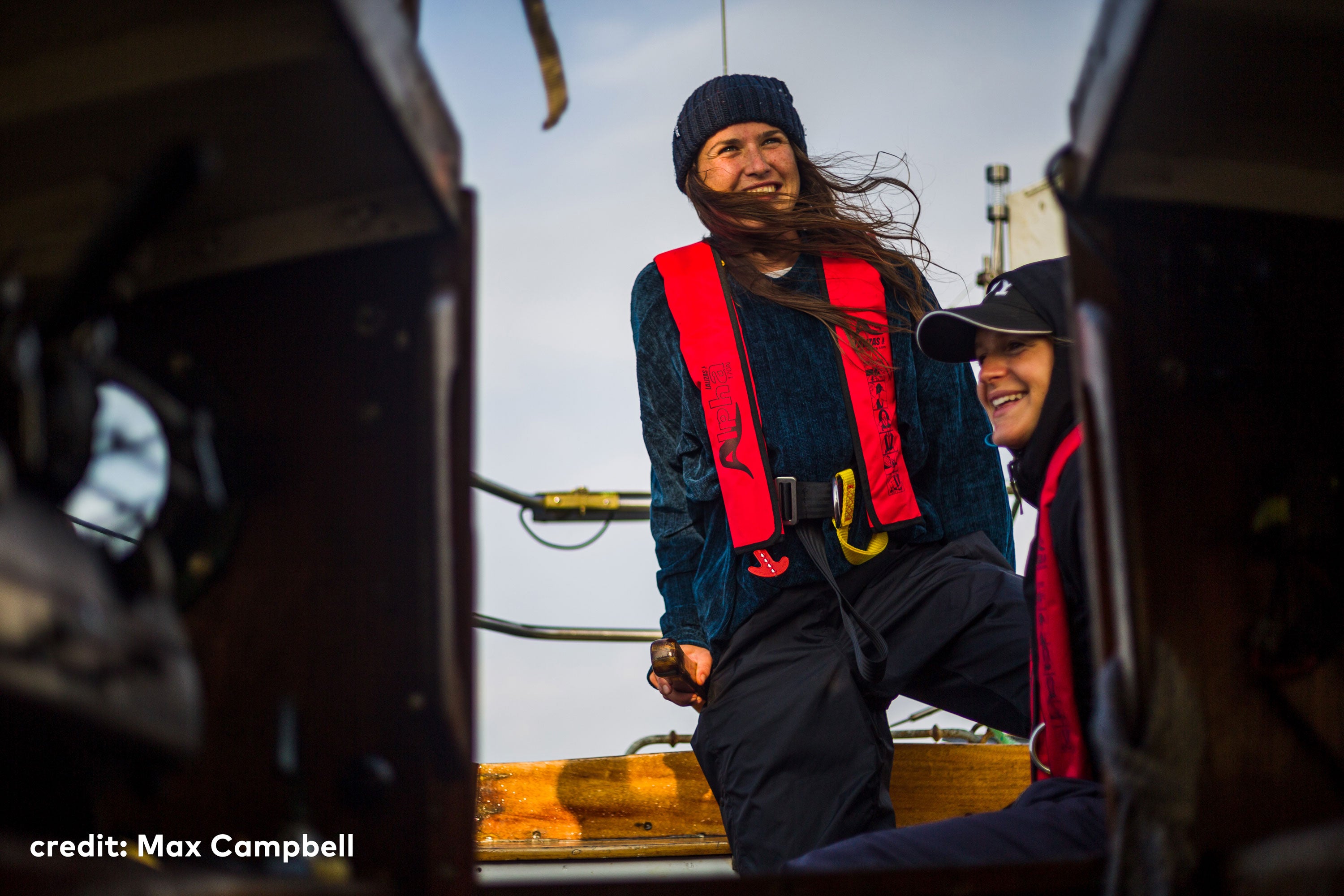 Two people sat on a boat, wearing cold weather clothing and red life jackets