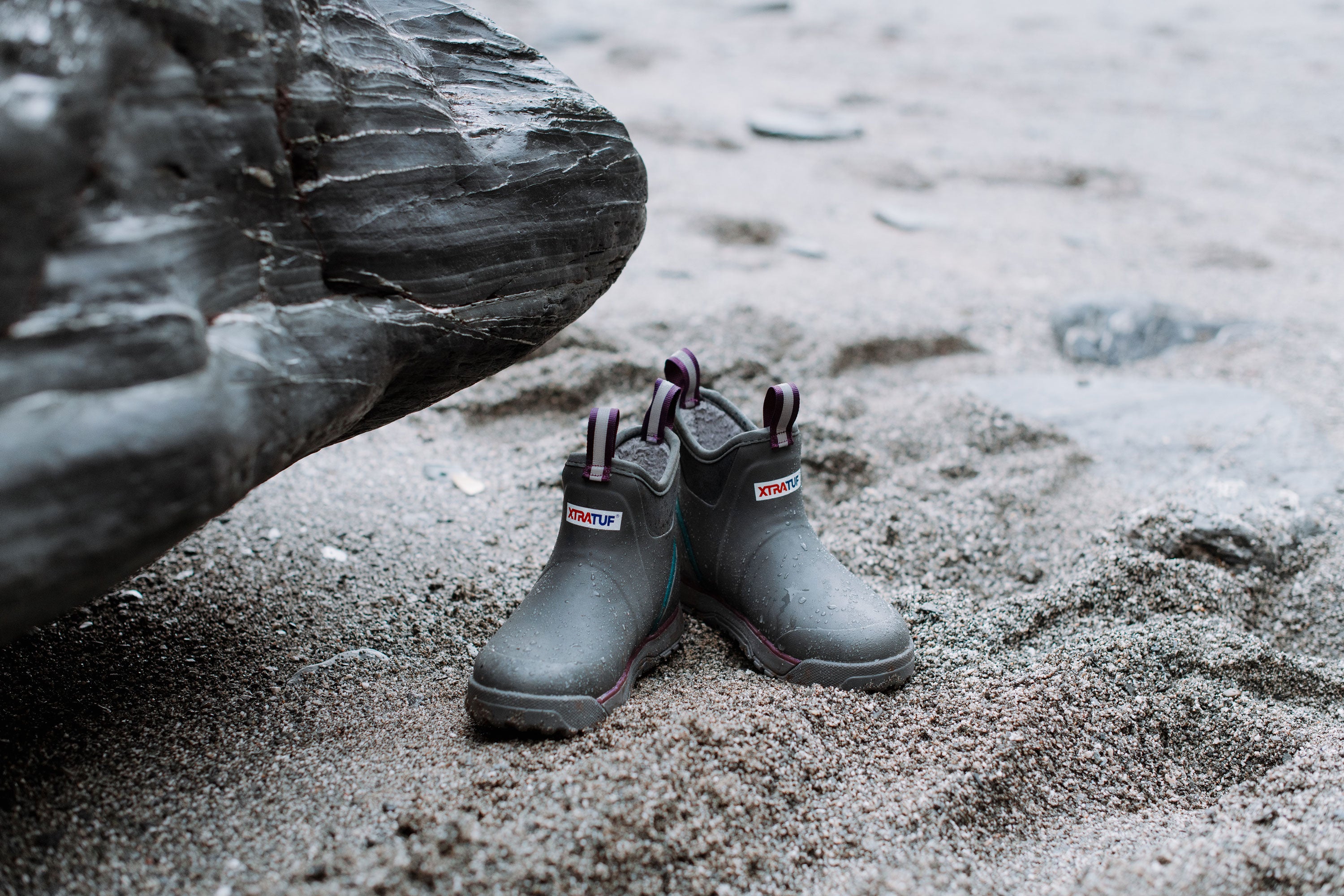 A pair of Xtratuf Ankle Deck Boots on a sandy beach, under a large rock