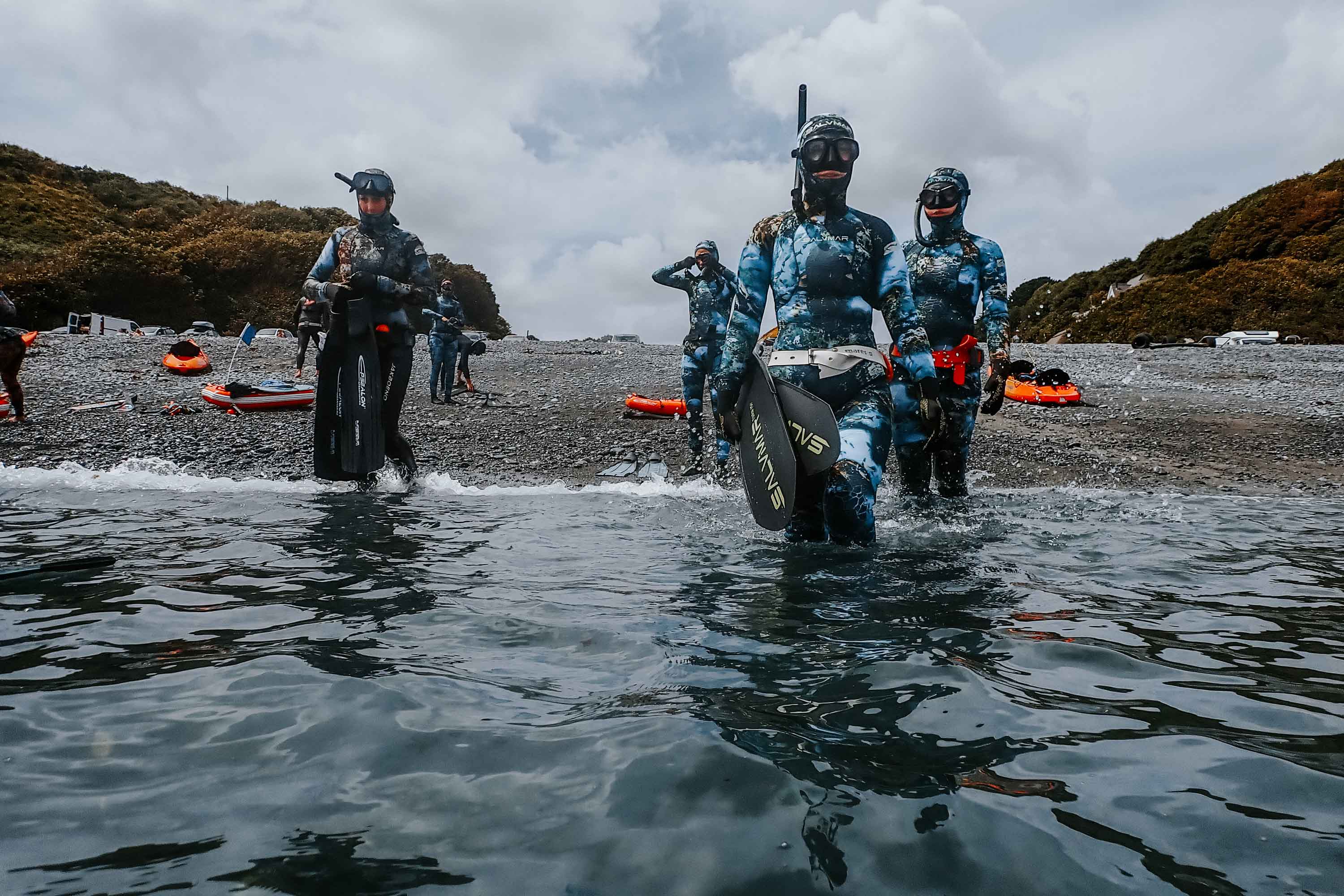 A group of people walking into water from a rocky beach, wearing diving and scuba gear