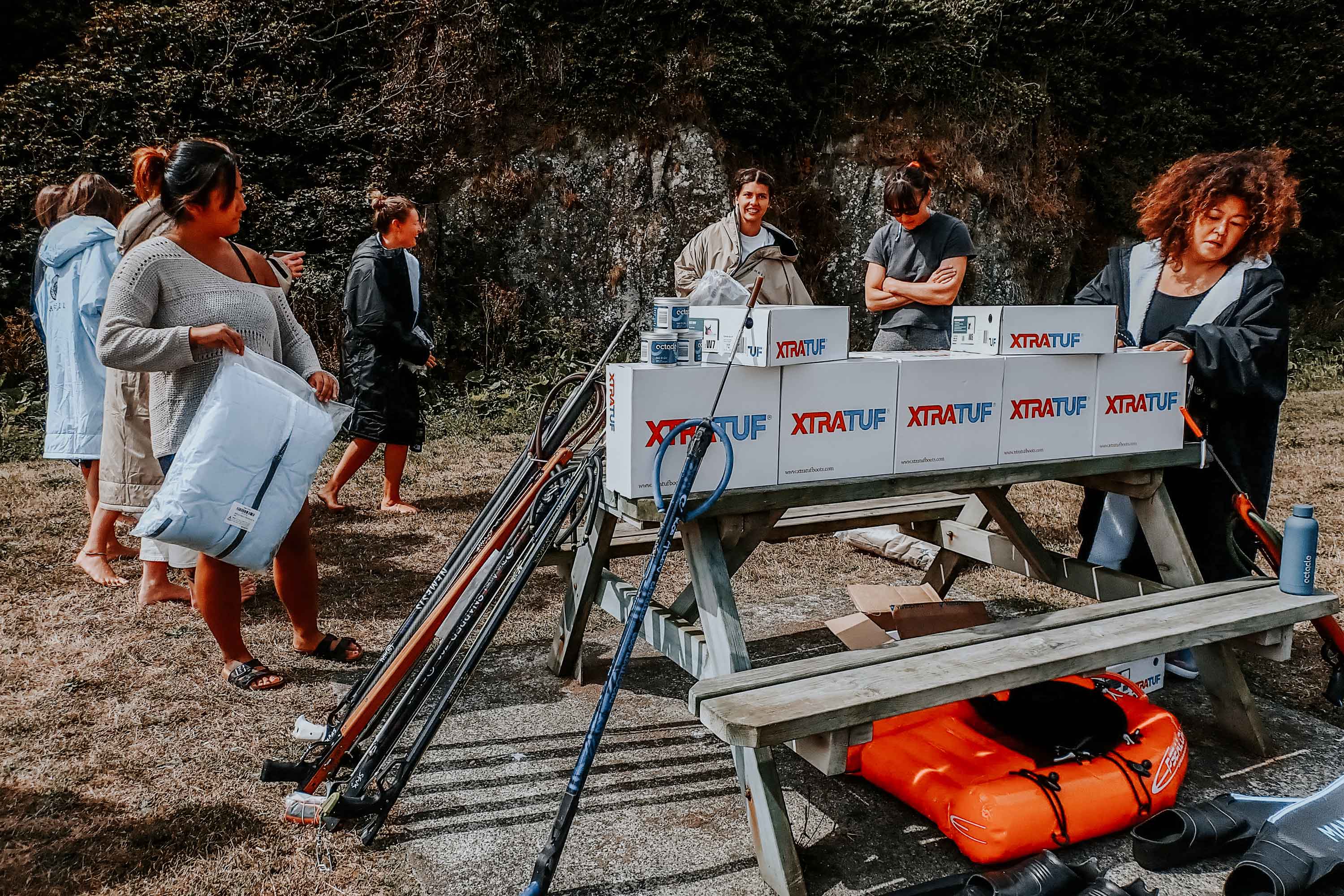 People on landing changing out of their scuba gear with spear guns propped up against a wooden bench with white Xtratuf boxes on top