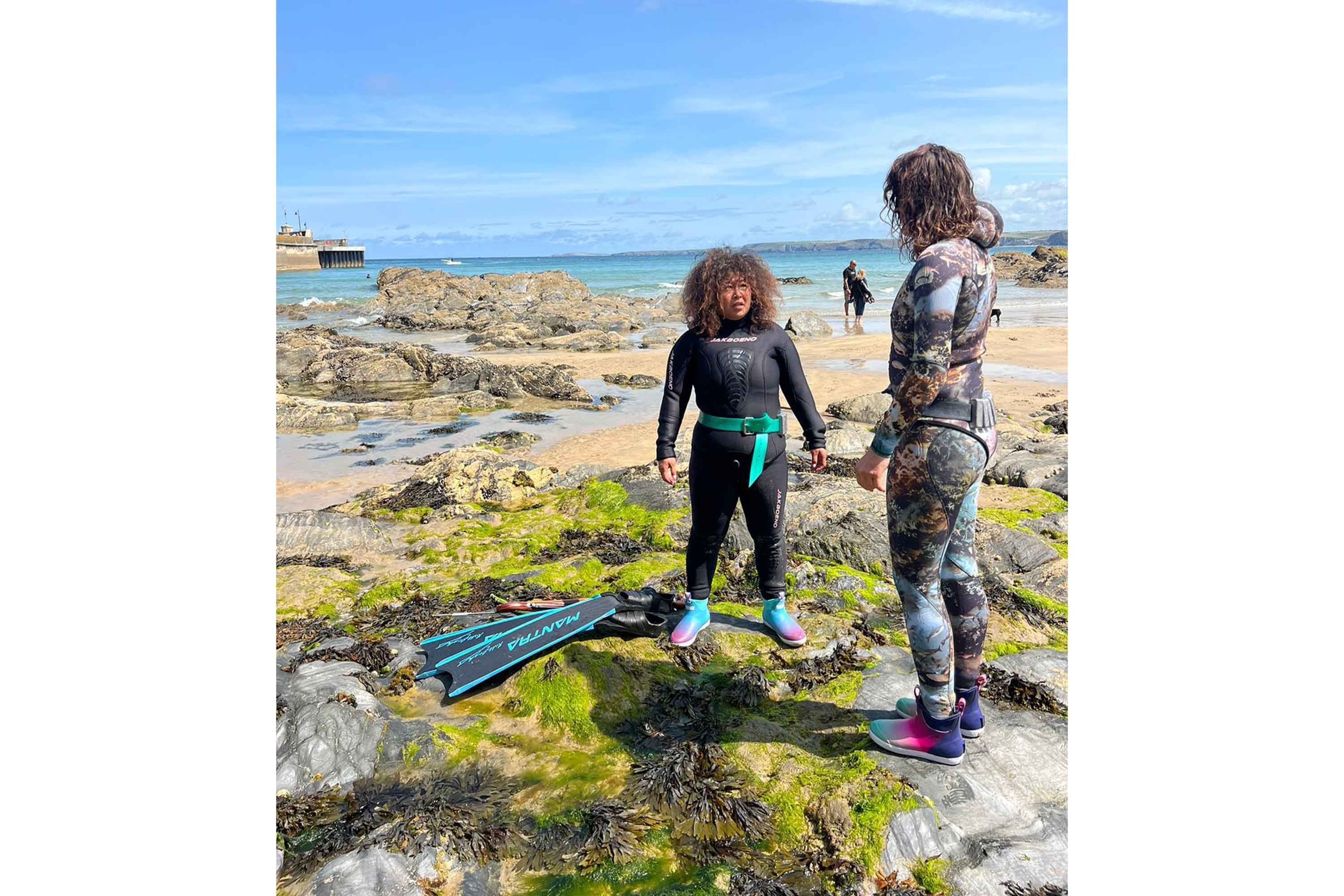Two women stood on seaweed covered rocks on a sunny beach, both wearing Xtratuf Aurora Ankle Deck Boots