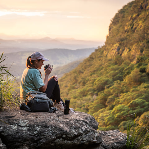 Woman drinking matcha or coffee sits on a rock overviewing a valley in Australia