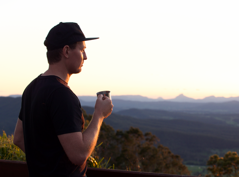 man holding cup of yerba mate tea whilst overlooking the sun setting over the mountains