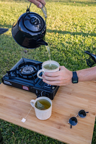 Black tea pot pouring Green Tea into camping mug with Australian background of grass, all featured on a wooden table with a camping cooker