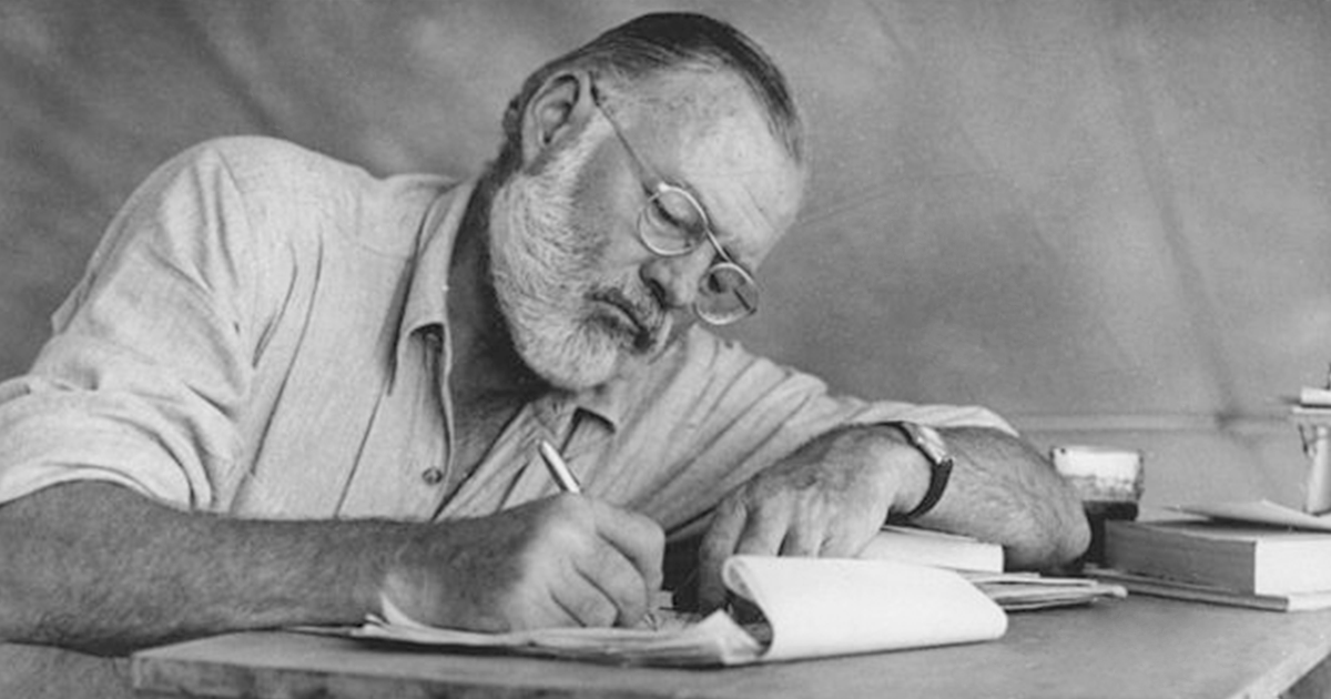 Photograph of Ernest Hemingway sitting at a table writing while at his campsite in Kenya, 1953.