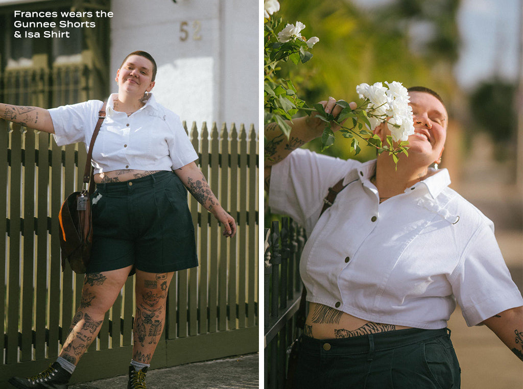 Frances poses against a picket fence and smells a white flower. They are wearing Gunnee Shorts in Green and an Isa Shirt in White.