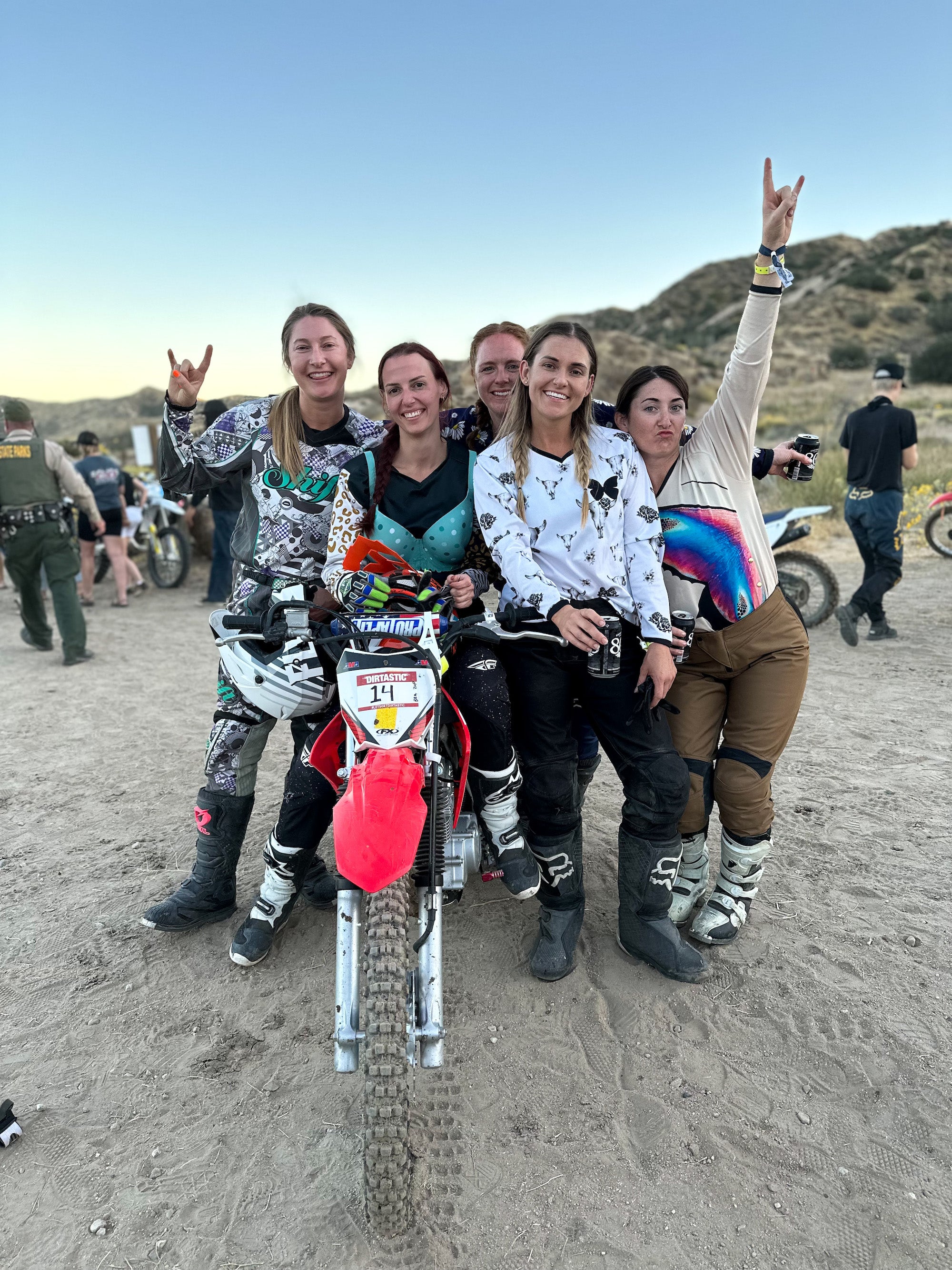 Five women gathered around a dirt bike, cheering after winning a relay race. They are wearing cute women's dirt bike gear by MCREY.