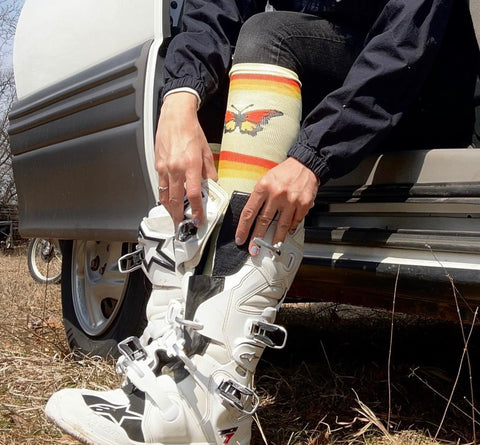 Woman wearing a retro yellow sock with a monarch butterfly on it and orange and yellow stripes. She is putting her foot in a white Alpinestars dirt bike boot. 